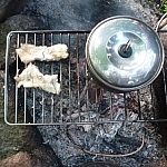 Two fillets next to a small backpacking pot, cooking on a campfire grill.
