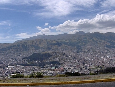 View of a city nestled in a valley, mountains in the backdrop and the shoulder of the highway visible in the foreground, depicting that the road is high up on a mountain above the city.