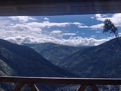 From Bellavista while hiking in Baños, we saw this view of mountains below a blue sky with white clouds, a wooden railing in the foreground.