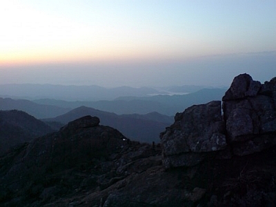 View from a mountain peak at sunrise, light mist hovering above the mountains, the sun piercing the cool blue morning sky in the distance.