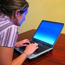 A woman sits at a table, typing on a laptop.
